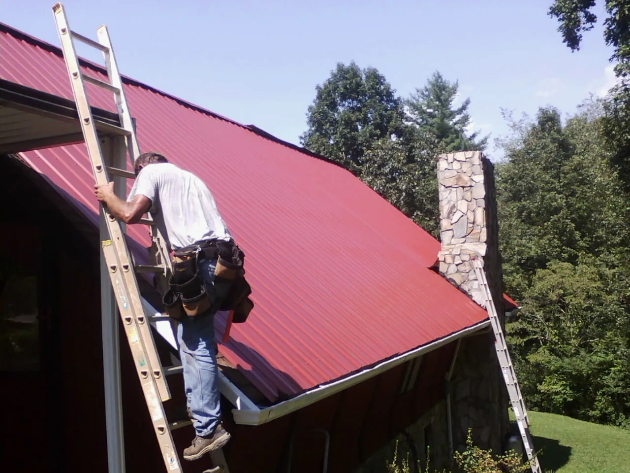 Roofer on a ladder beside a red metal roof and stone chimney — Soffit & Fascia Repair by Roofing Brothers in Yorktown